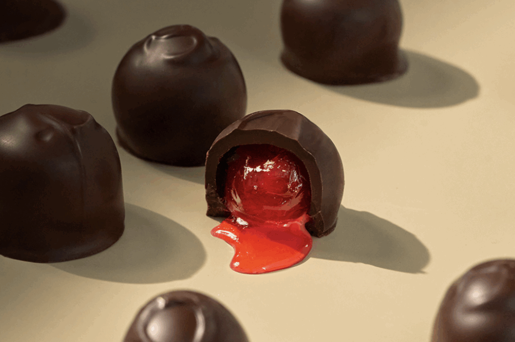 A photo of a cream coloured table top, featuring an up close image of seven Cordial Cherries, spread out at random. In the centre of the image, the featured Cordial Cherry chocolate is cut open, so we see the red Maraschino cherry contained within, with red cordial spreading out from around the cherry, onto the tabletop.